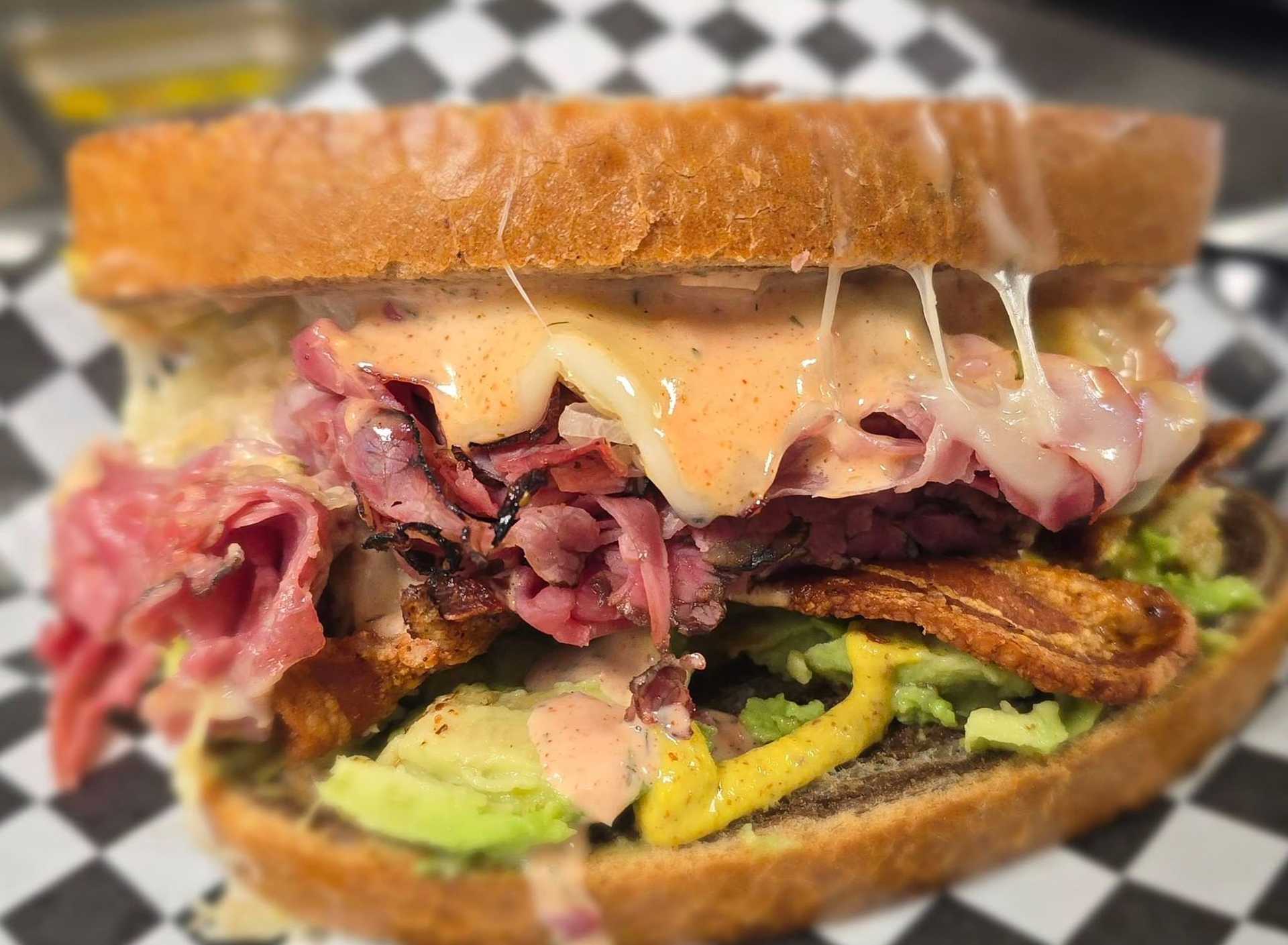 Sandwich with pastrami, avocado, cheese, and mustard on checkered paper.